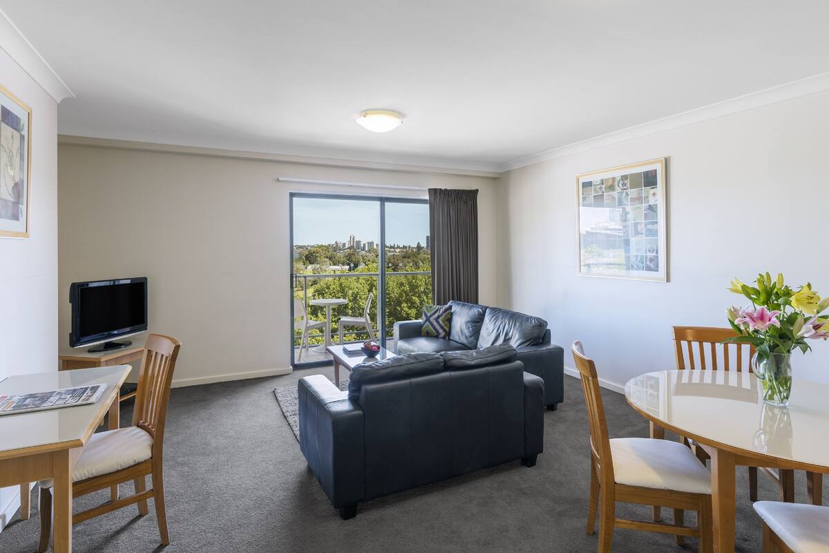 A modern living room with a sofa, a dining table, and chairs. There is a television on a stand, a large window with a view of greenery and buildings, and a framed picture on the wall.