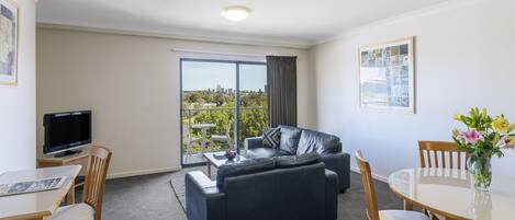 A modern living room with a sofa, a dining table, and chairs. There is a television on a stand, a large window with a view of greenery and buildings, and a framed picture on the wall.