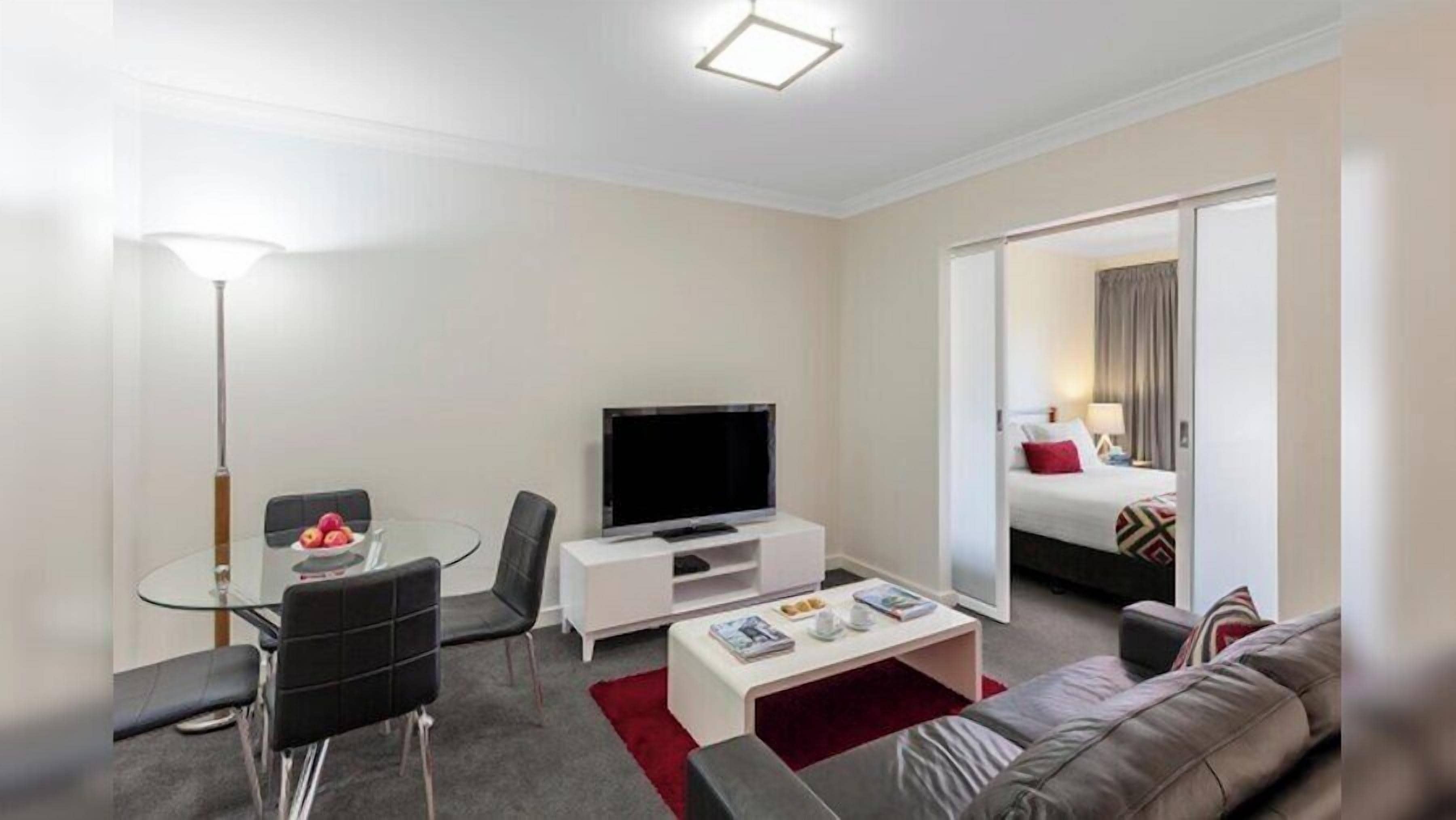 A modern hotel room with a flat-screen TV, a glass dining table, a sofa, and a bed with a red pillow.