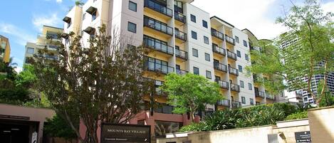 A multi-story residential building with balconies, a sign indicating amenities like a courtyard restaurant and car parks, and lush greenery around the property.