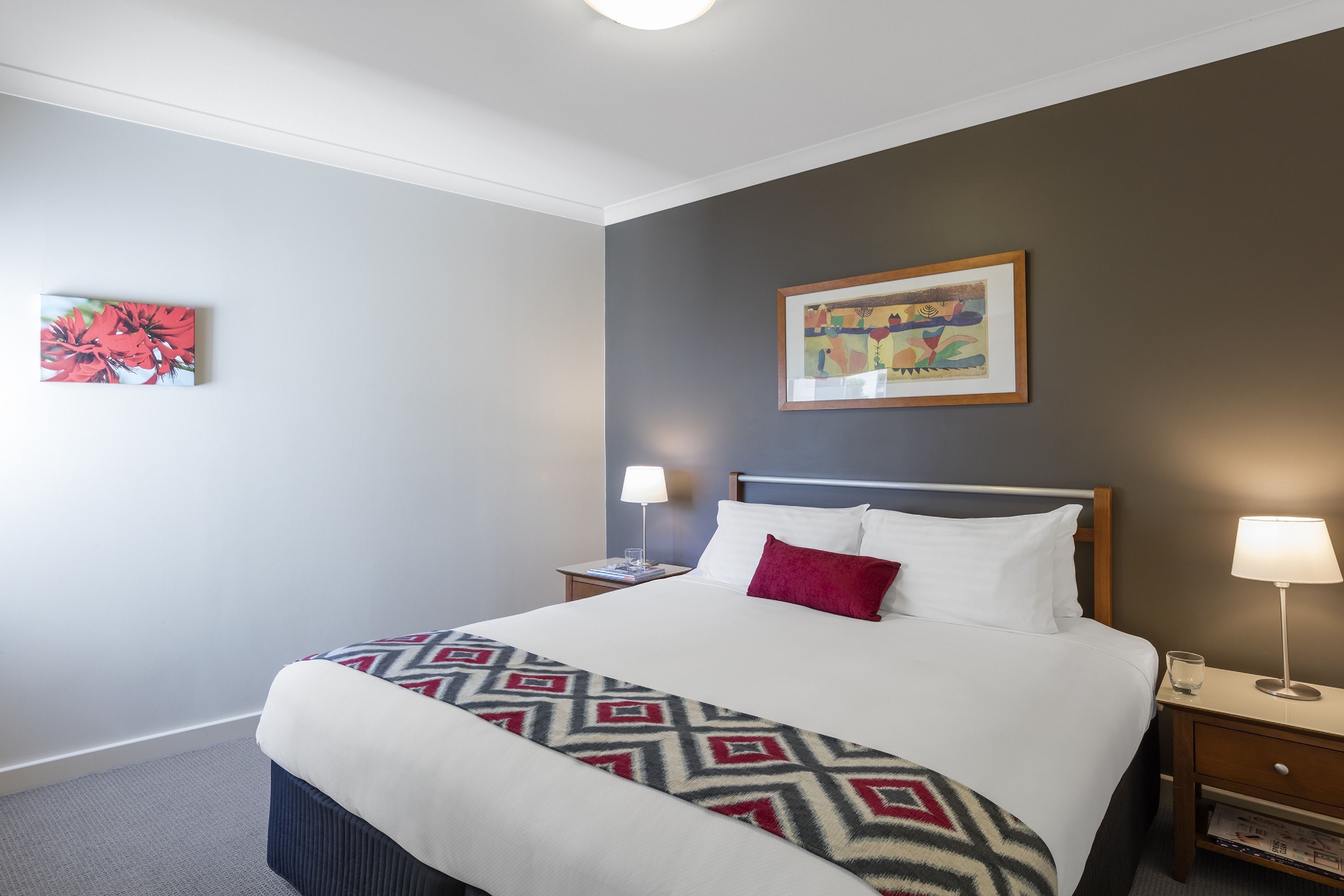A neatly made bed with a patterned throw, two bedside lamps, a framed artwork above the bed, and a small table with a lamp and glass.
