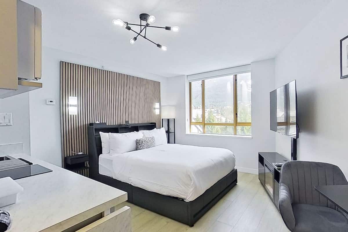 Una habitación de hotel moderna con cama, escritorio, silla, televisor y un ventanal grande.