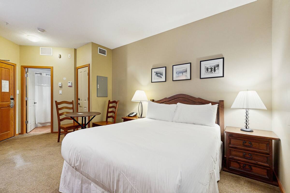 Habitación de hotel con una cama grande, dos mesitas de noche con lámparas, un aparador de madera, una silla, una pequeña mesa y una puerta que da a un baño.