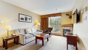 A cozy living room with a sofa, armchair, and coffee table, a fireplace, and a large window with a view of trees. - Whistler Cascade Lodge (Whistler)