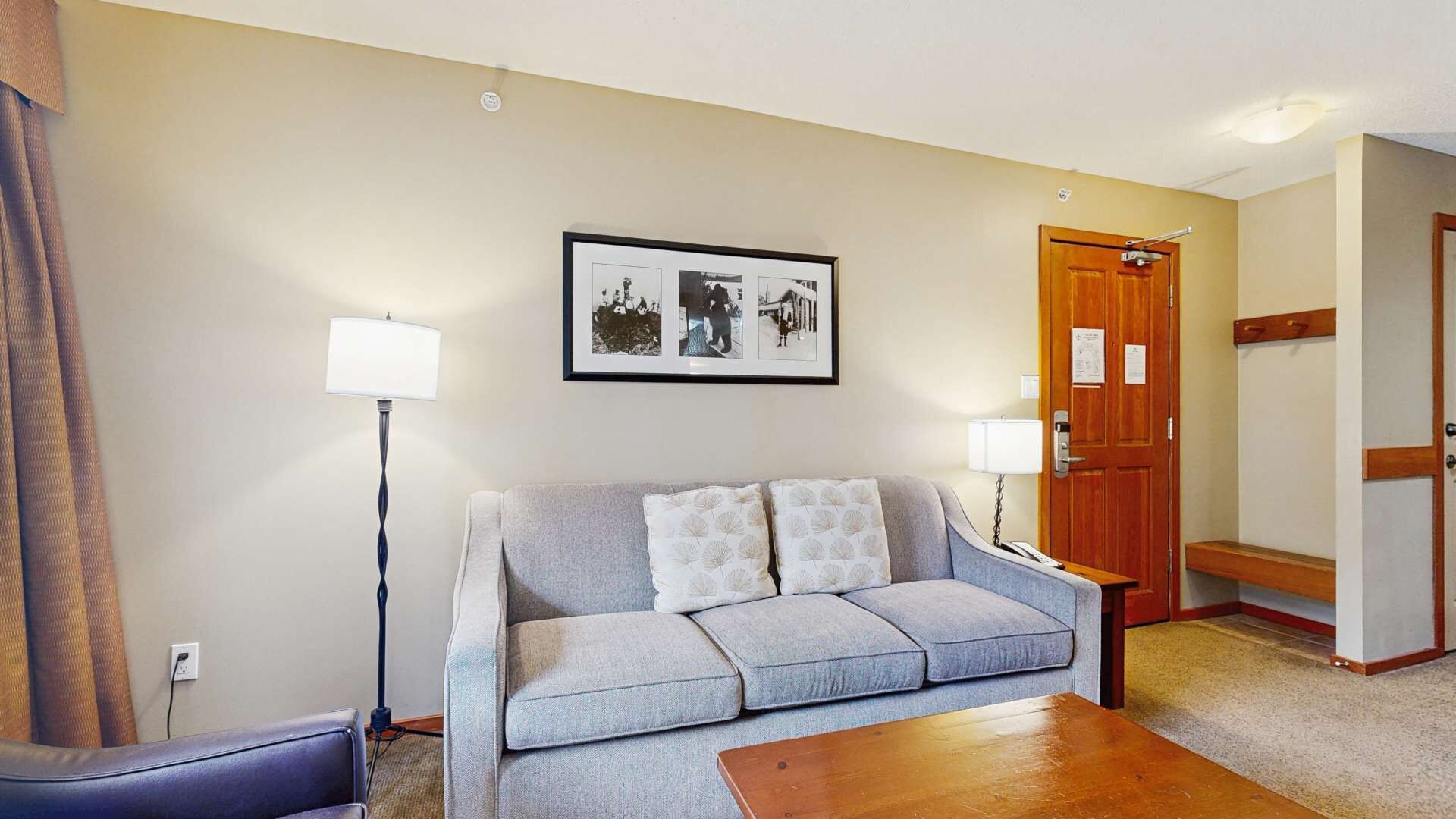A living room with a grey sofa, two white pillows, a wooden coffee table, a floor lamp, and a framed photo on the wall.