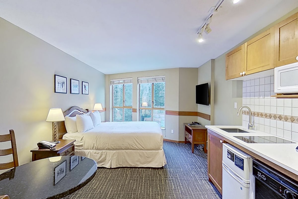 Habitación de hotel con cama, mesita de noche, lámpara, televisión y kitchenette.
