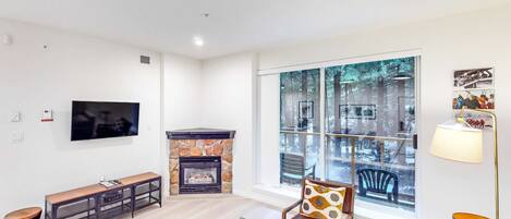 A modern living room with a stone fireplace, a flat-screen TV, and a large window offering a view of trees.