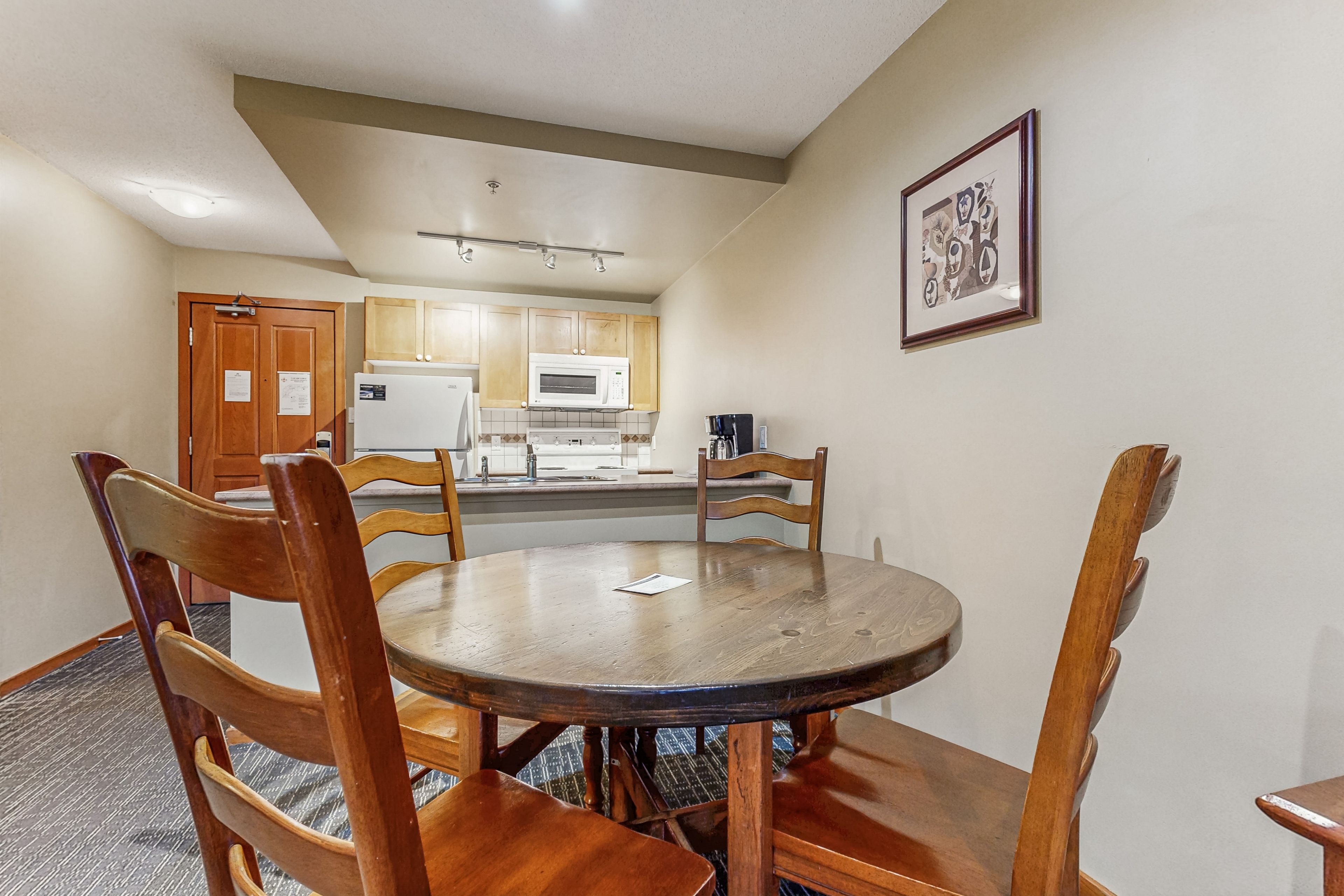 A dining area with a round wooden table and chairs, a kitchen in the background, and a framed picture on the wall.