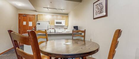 A dining area with a round wooden table and chairs, a kitchen in the background, and a framed picture on the wall.
