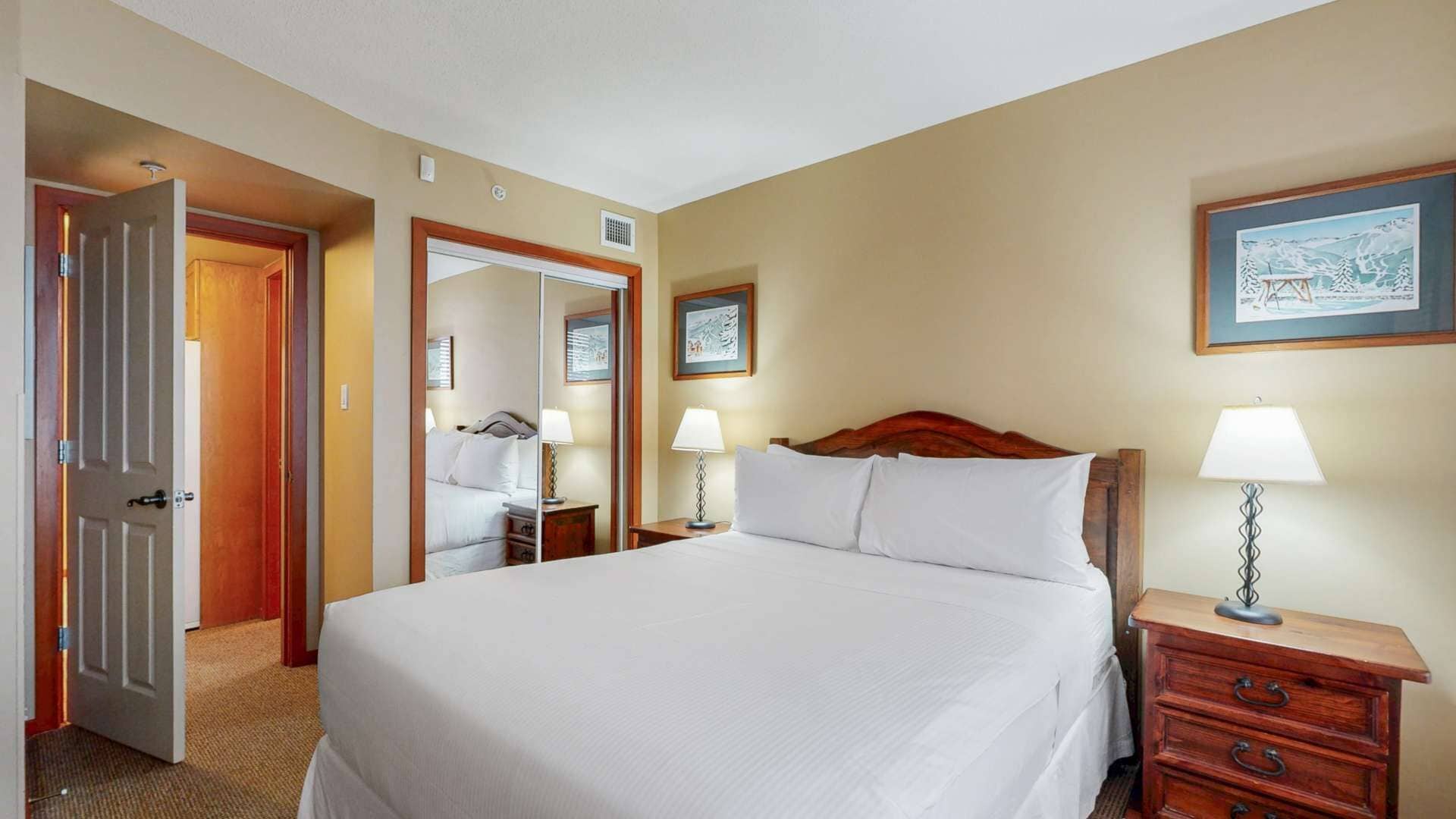Hotellihuone, jossa on suuri sänky, kaksi yöpöytää valaisimineen, puinen sängynpääty, ovi ja peili.