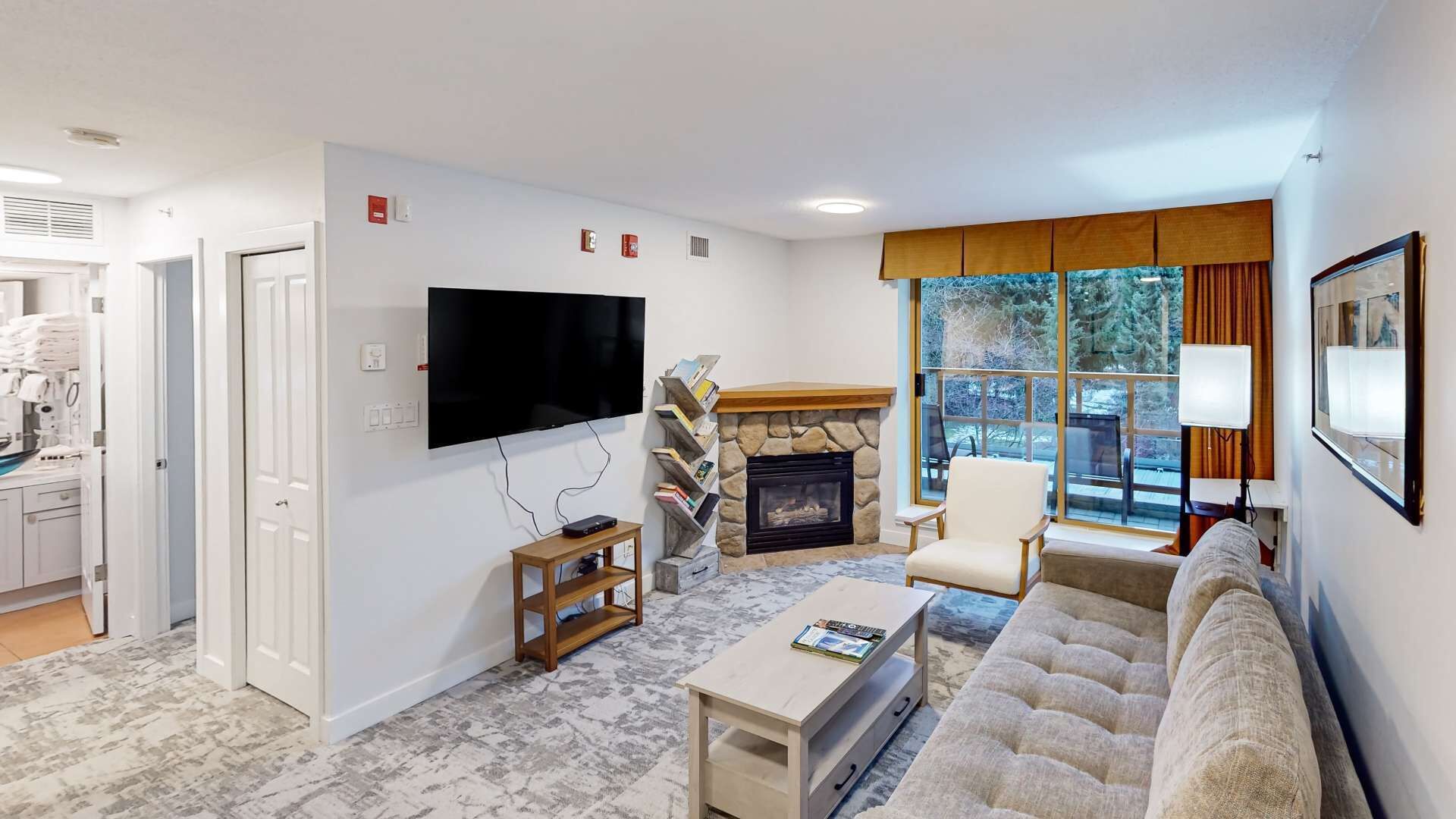 A modern living room with a flat-screen TV, a stone fireplace, a comfortable sofa, and a large window offering a view of trees.