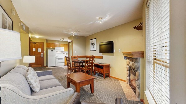 A living room with a sofa, coffee table, and TV. There is a kitchen area with a fridge and microwave. A fireplace is visible, and the room has a ceiling fan.