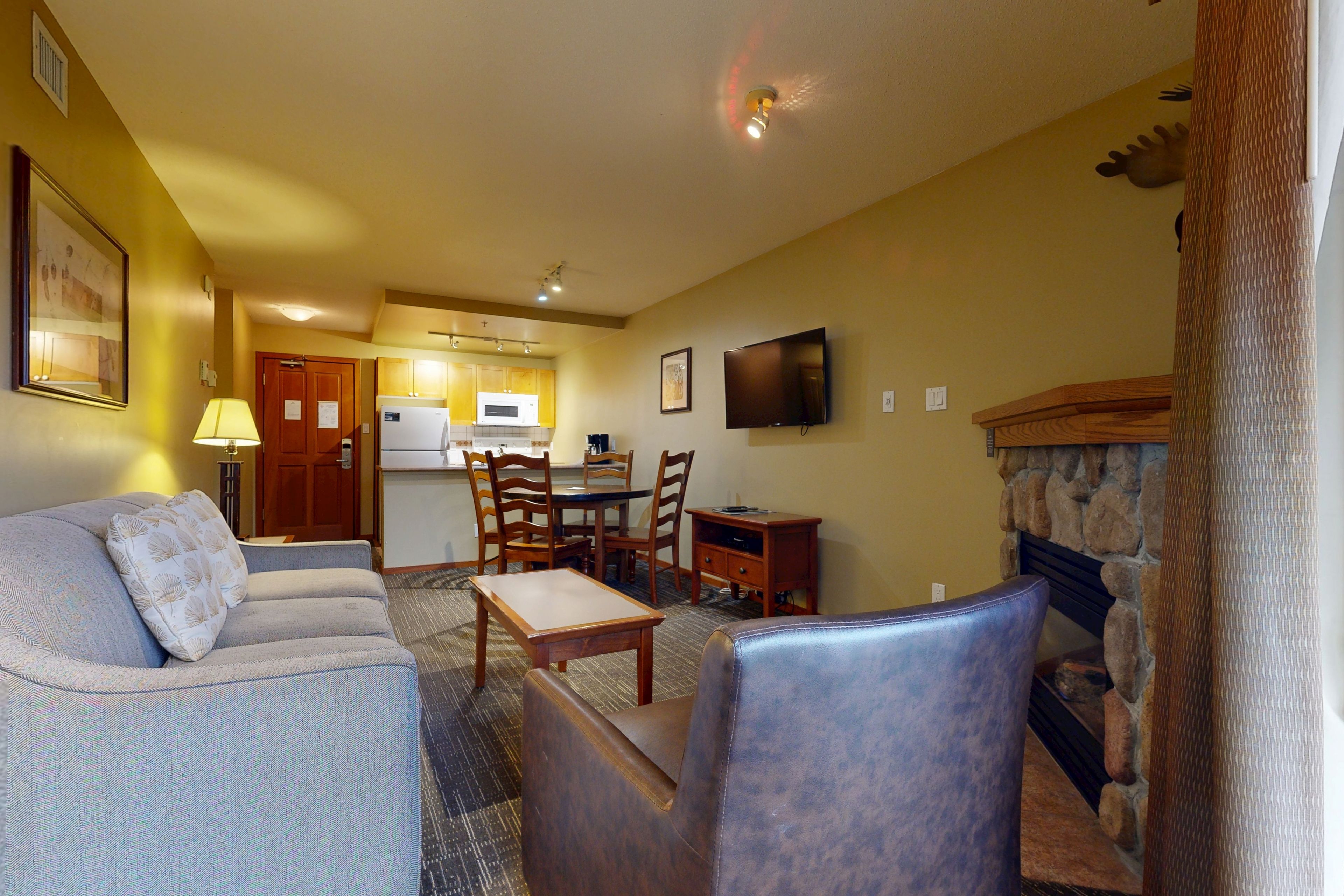 A living room with a sofa, armchair, and a fireplace. There is a dining table with chairs and a kitchen in the background.