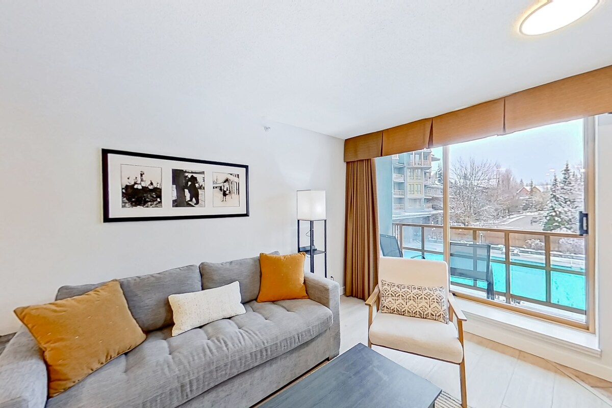 Una sala de estar moderna con un sofá gris, una mesa de centro y un sillón con un cojín estampado. Hay un ventanal con vista a una piscina y edificios.