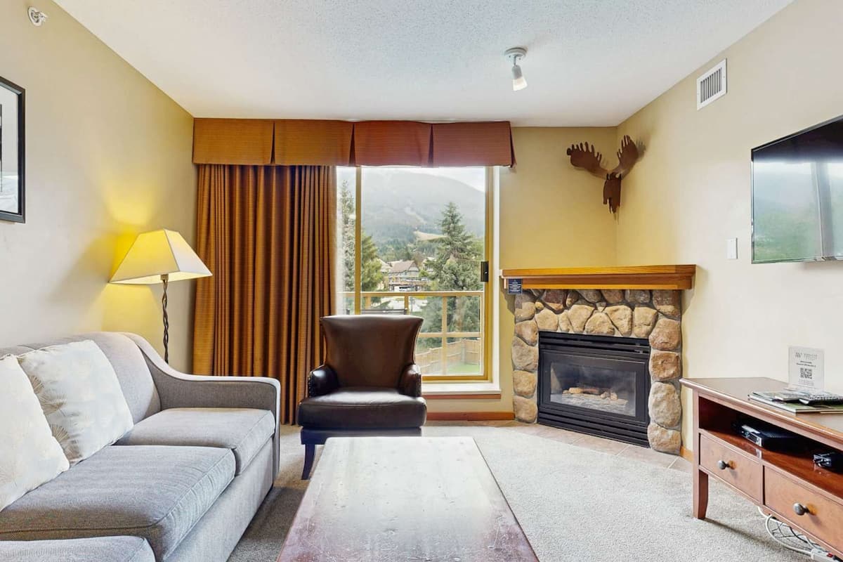 Una sala de estar con sofá, sillón, chimenea y un ventanal con cortinas.