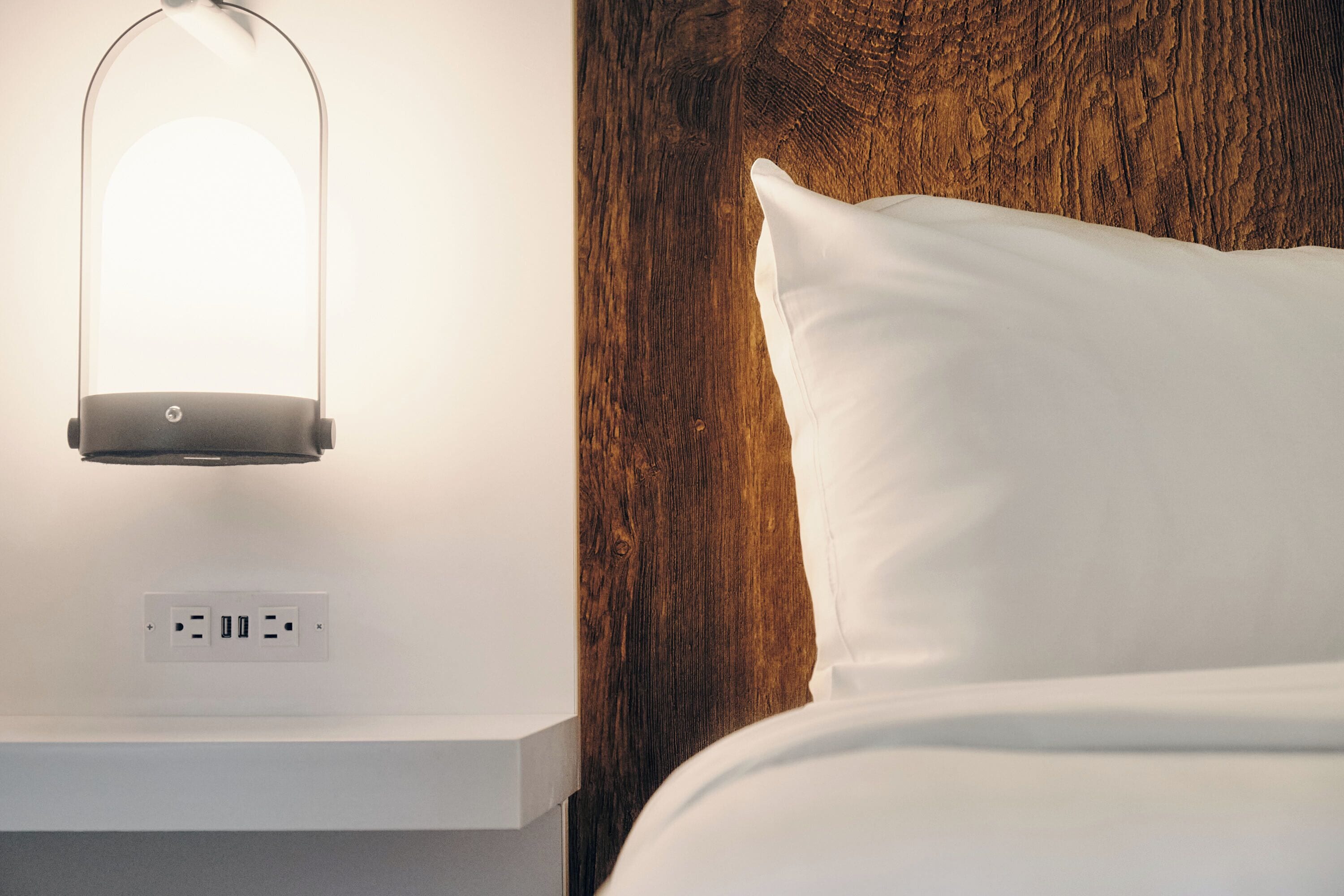A neatly made bed with a white pillow and a wooden headboard. Below the bed, there is a white nightstand with a lamp and a power outlet.