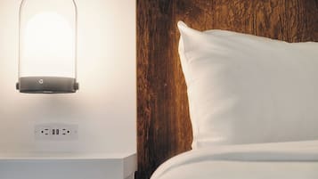 A neatly made bed with a white pillow and a wooden headboard. Below the bed, there is a white nightstand with a lamp and a power outlet.