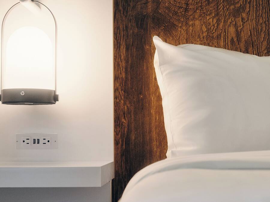 Una cama bien hecha con un almohadón blanco y un cabecero de madera. Debajo de la cama, se encuentra una mesita de noche blanca con una lámpara y un enchufe.