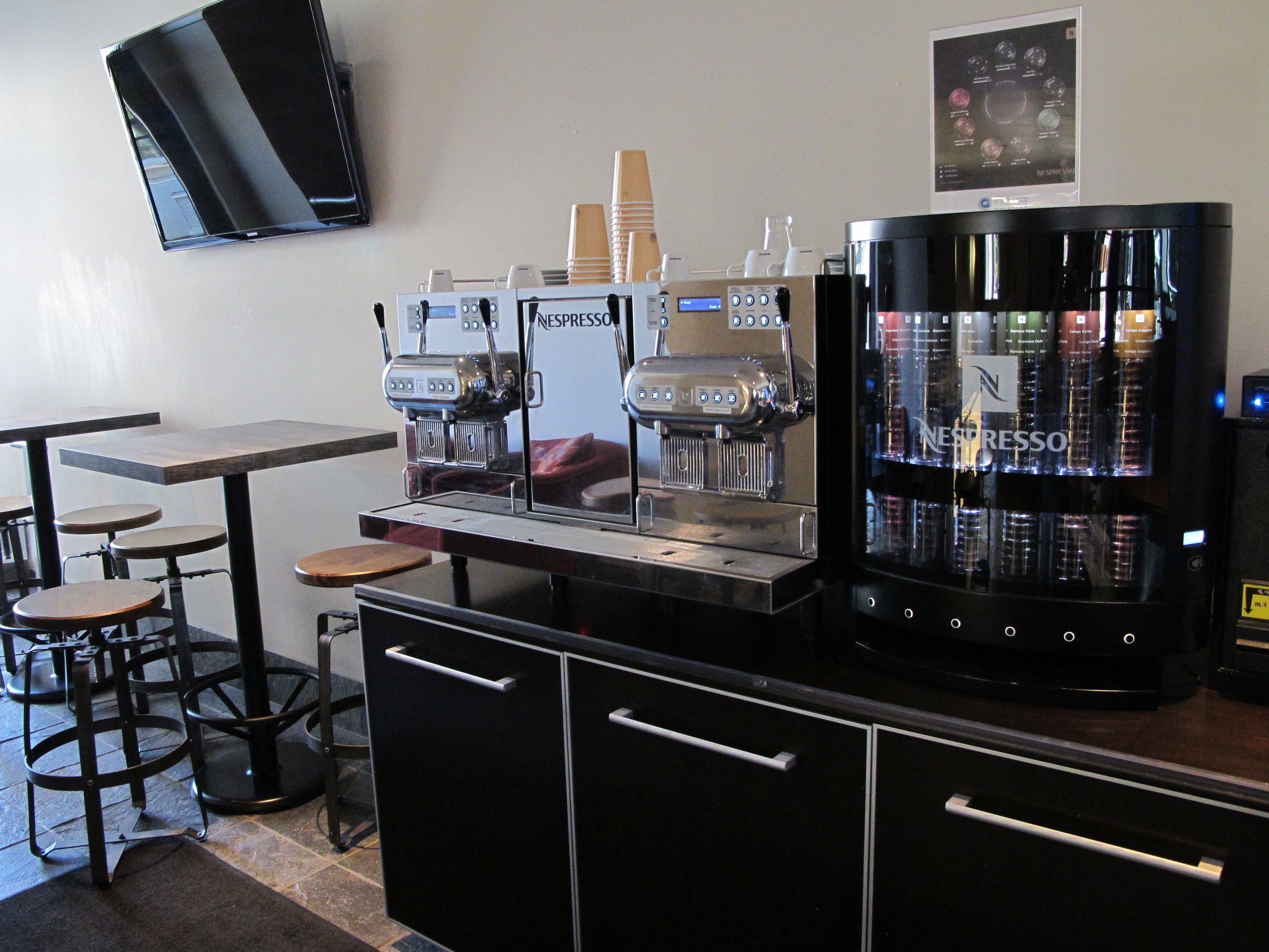 Una cafetería con dos máquinas de espresso, un molinillo de café y un refrigerador lleno de productos Nespresso. Hay taburetes y un televisor de pantalla plana montado en la pared.