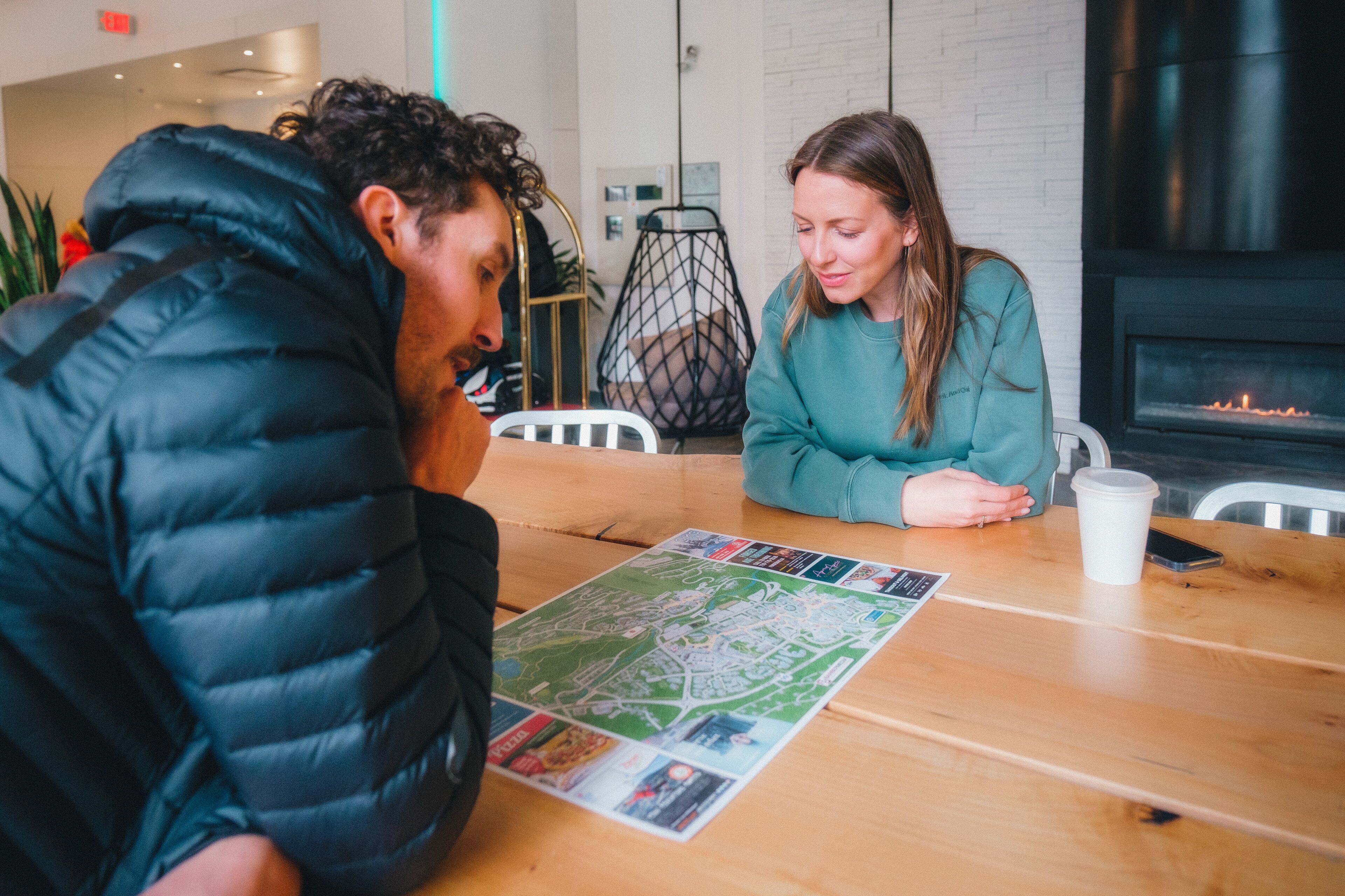 Two people sitting at a wooden table with a map, a white cup, and a smartphone.
