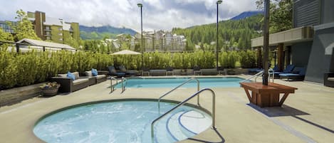 Zona de piscina con sillones de descanso, una mesa de madera y vistas a montañas.