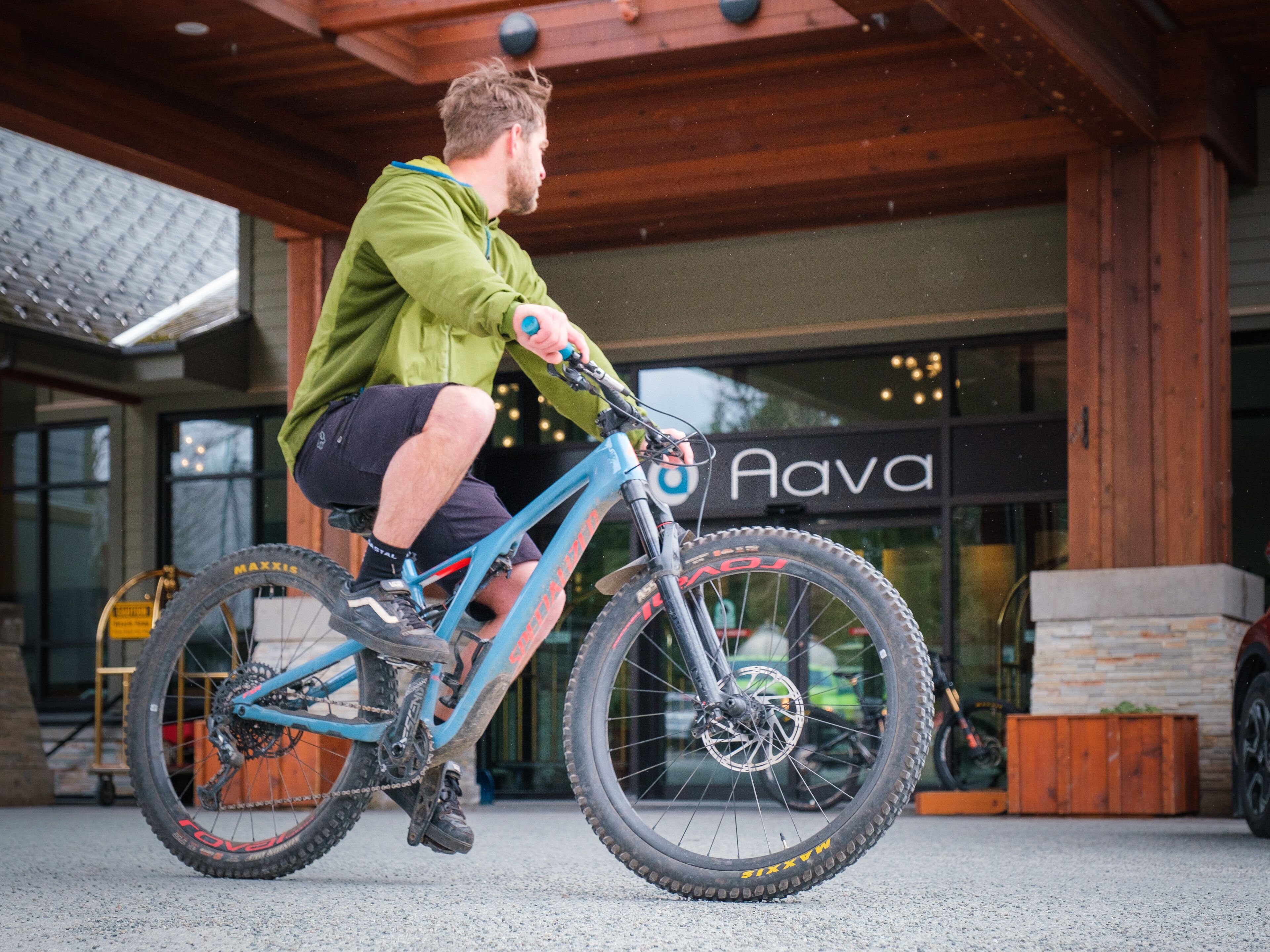 A person riding a blue mountain bike in front of a building with a sign that reads "Aava".