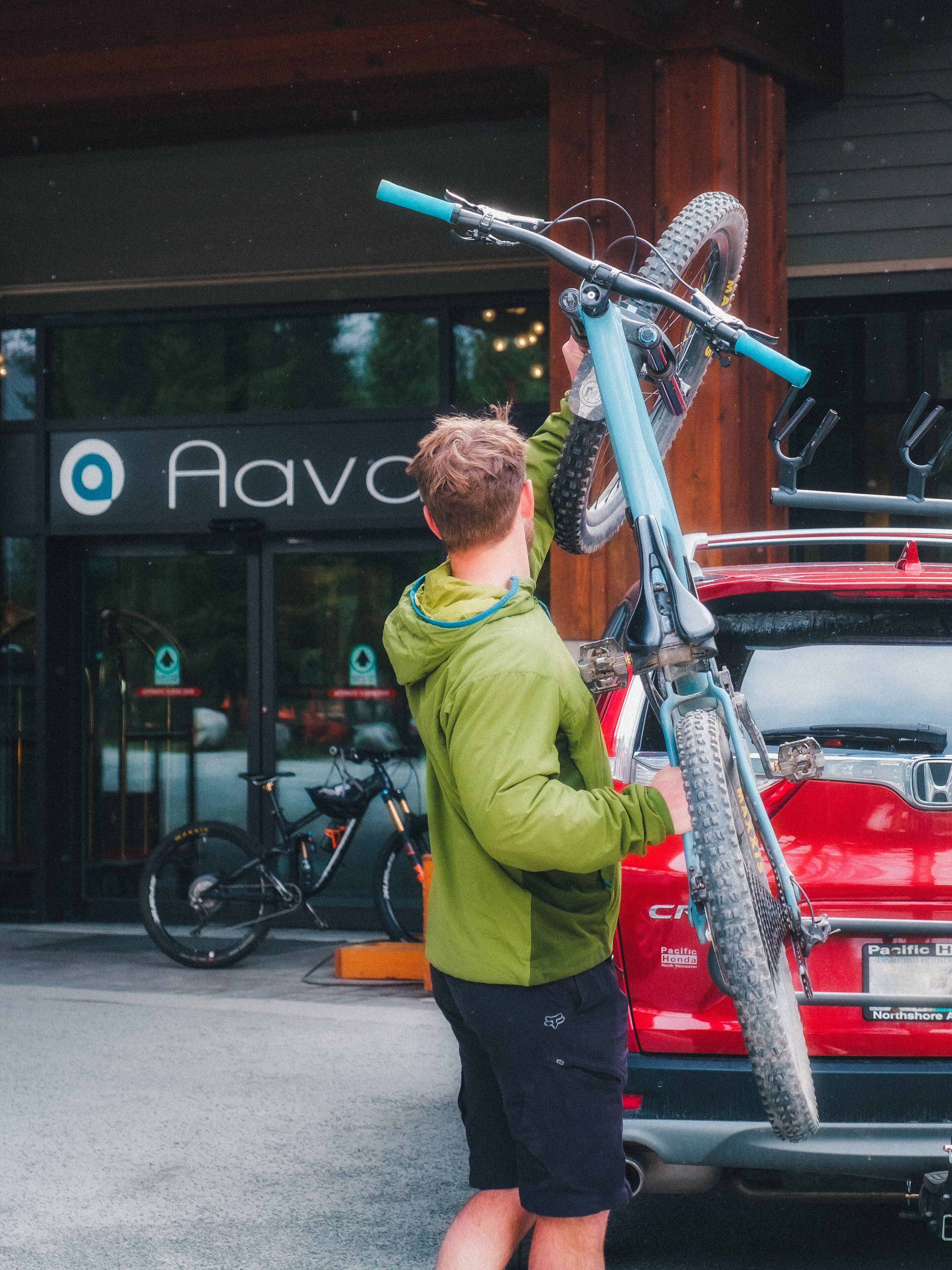 Une personne qui porte un vélo au-dessus d’une voiture rouge devant un bâtiment sur lequel on peut lire l’inscription « Ava ».
