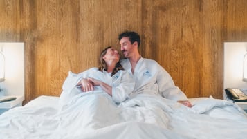 A couple in white robes on a bed with a wooden headboard, a bedside phone, and a lamp on a nightstand.