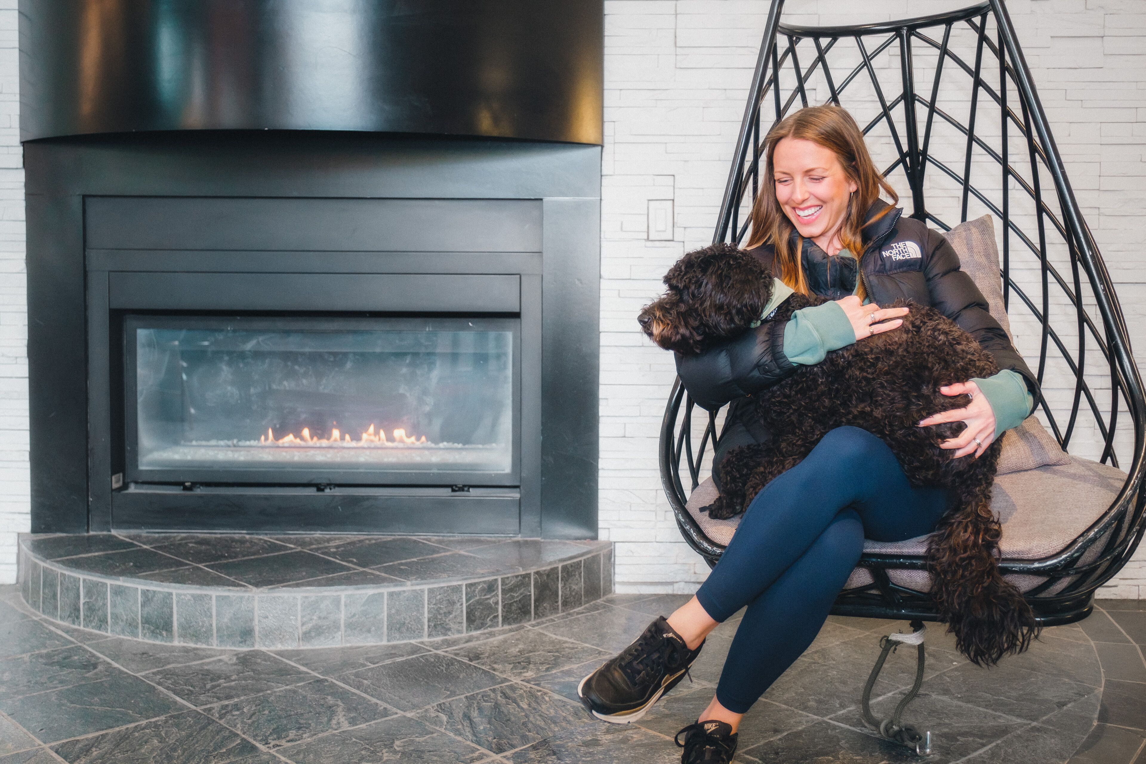 A person sitting in a black chair by a fireplace with a dog.