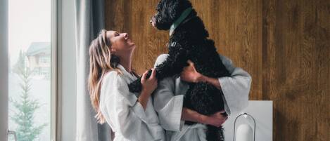 Een persoon in een witte gewaad die een zwarte hond vasthoudt in een kamer met een houten wand en een raam.