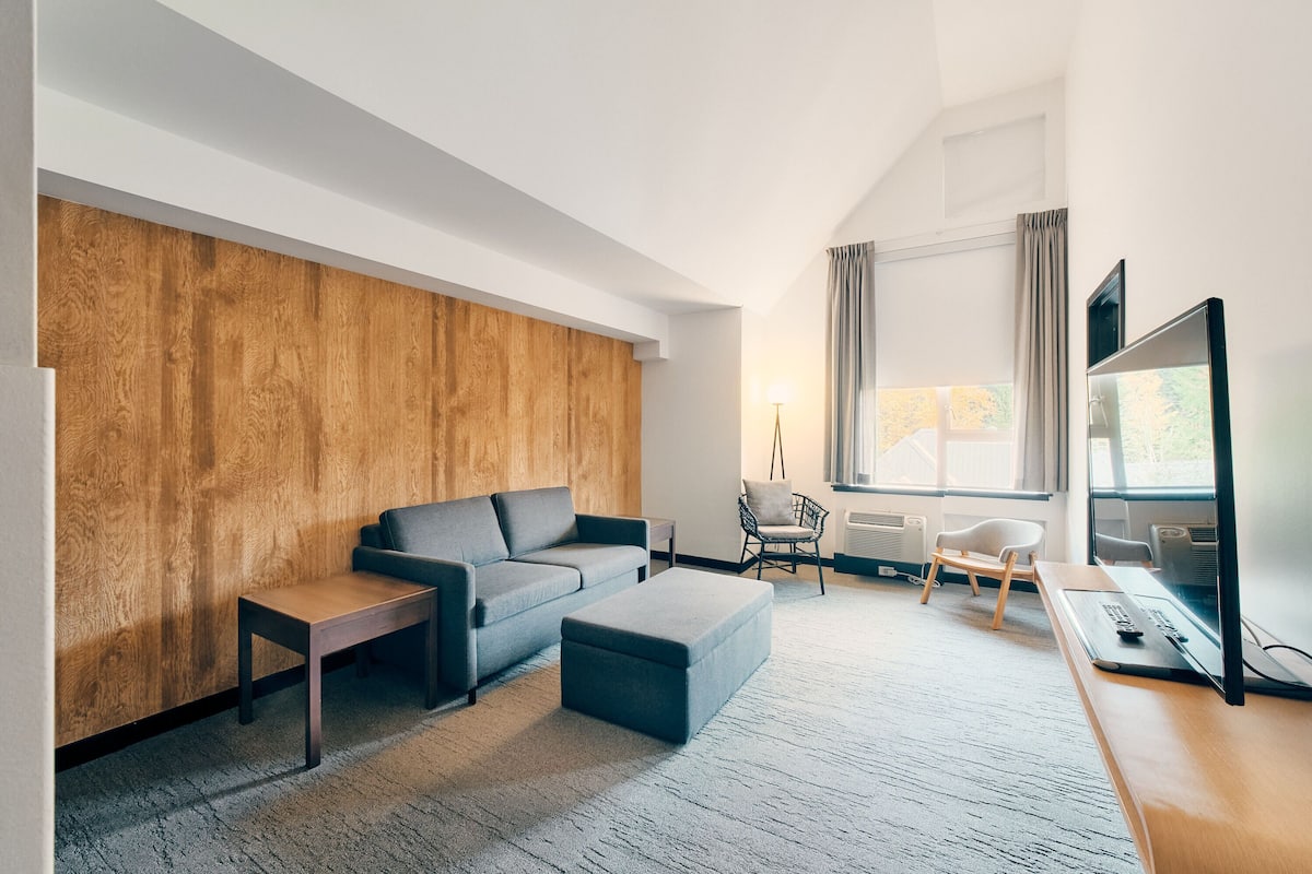 Una sala de estar moderna con un sofá gris, un muro decorativo de madera, una pequeña mesa, un otoman gris, un escritorio con una computadora y un televisor.