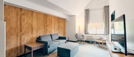 Una sala de estar moderna con un sofá gris, un muro decorativo de madera, una pequeña mesa, un otoman gris, un escritorio con una computadora y un televisor.