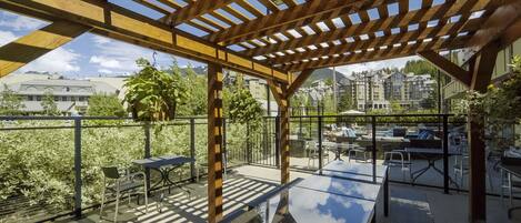 A covered outdoor seating area with tables and chairs, overlooking a cityscape with buildings and greenery.