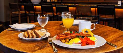 Un desayuno con platos de huevos, tocino, tostadas y fruta, junto con vasos de agua y zumo de naranja.