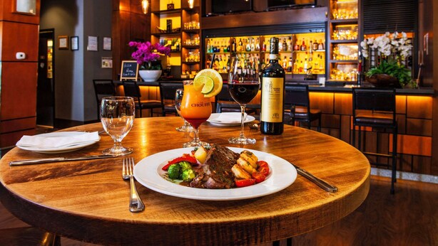 A round wooden table set with a meal, wine glasses, and a bottle of wine in a bar setting. - Grand Park Hotel Vancouver Airport, an Ascend Collection Hotel (Richmond)