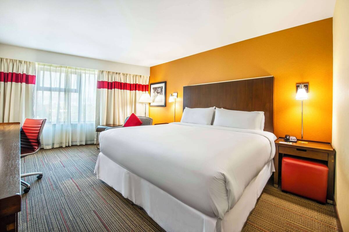 A hotel room with a large bed, a desk, a chair, a nightstand, and a lamp.