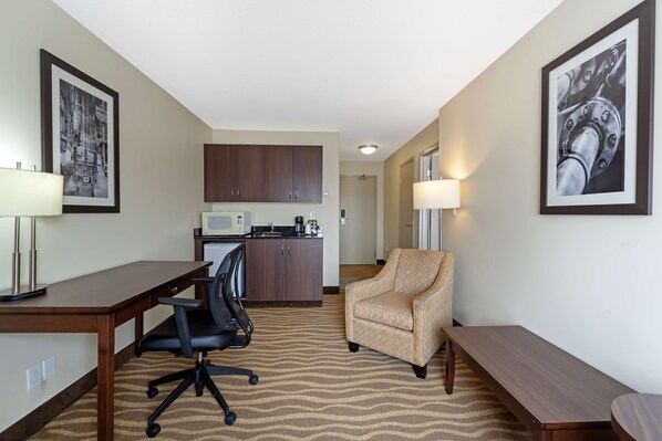 A hotel room with a kitchenette, a desk with a chair, a patterned armchair, and a bench.