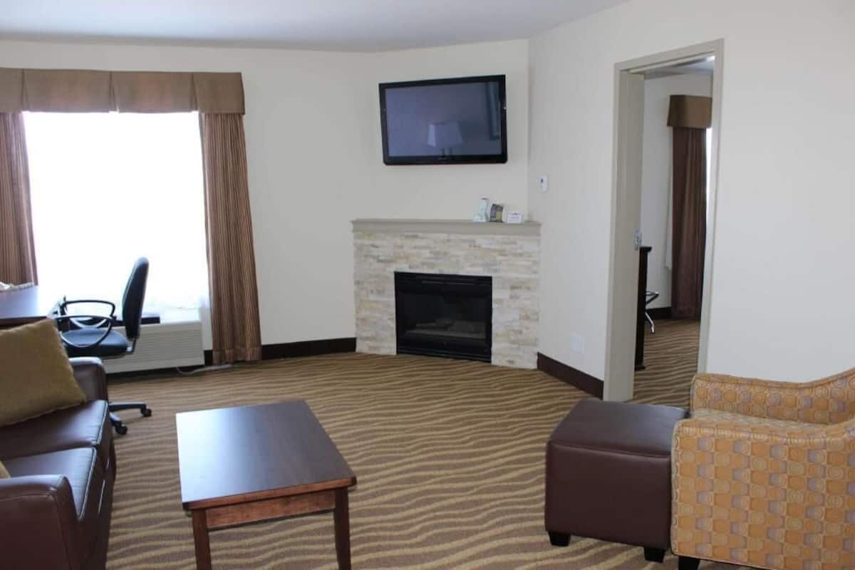 Habitación de hotel con chimenea, televisor montado en la pared, sofá, reposapiés y suelo alfombrado.