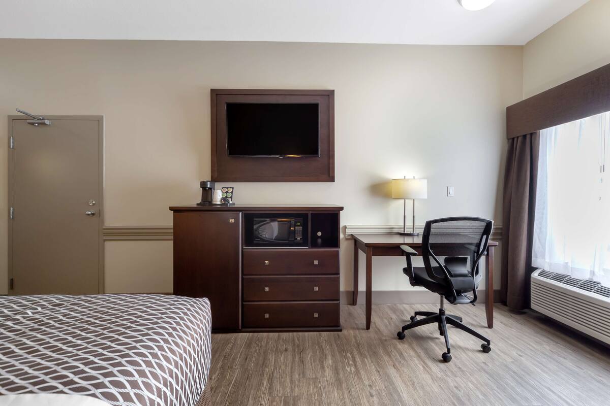 Habitación de hotel con cama, escritorio con silla, televisor montado en la pared y ventana con cortinas.