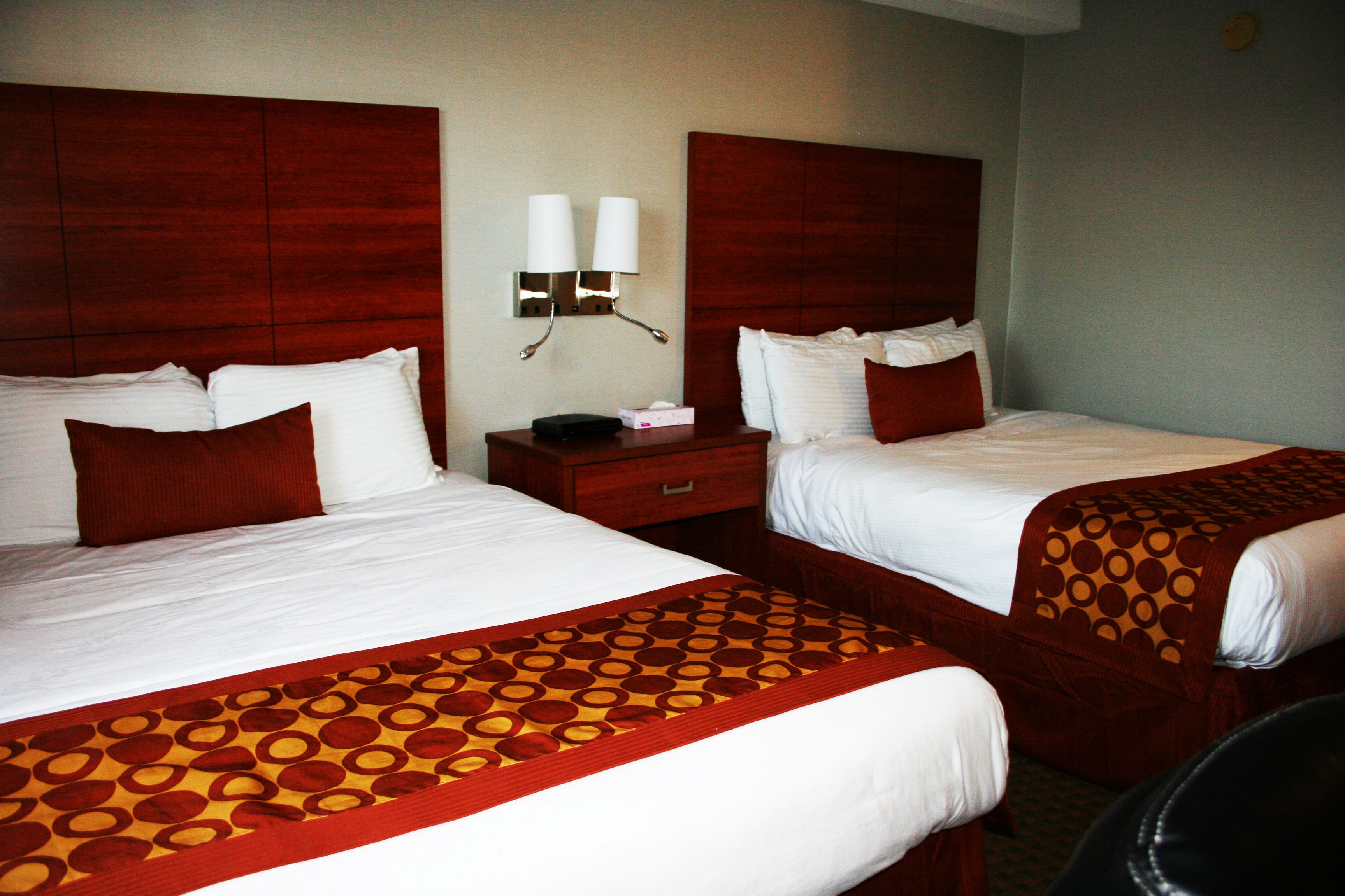 Dos camas de hotel juntas con sábanas blancas, un cabecero de madera y una colcha roja estampada.