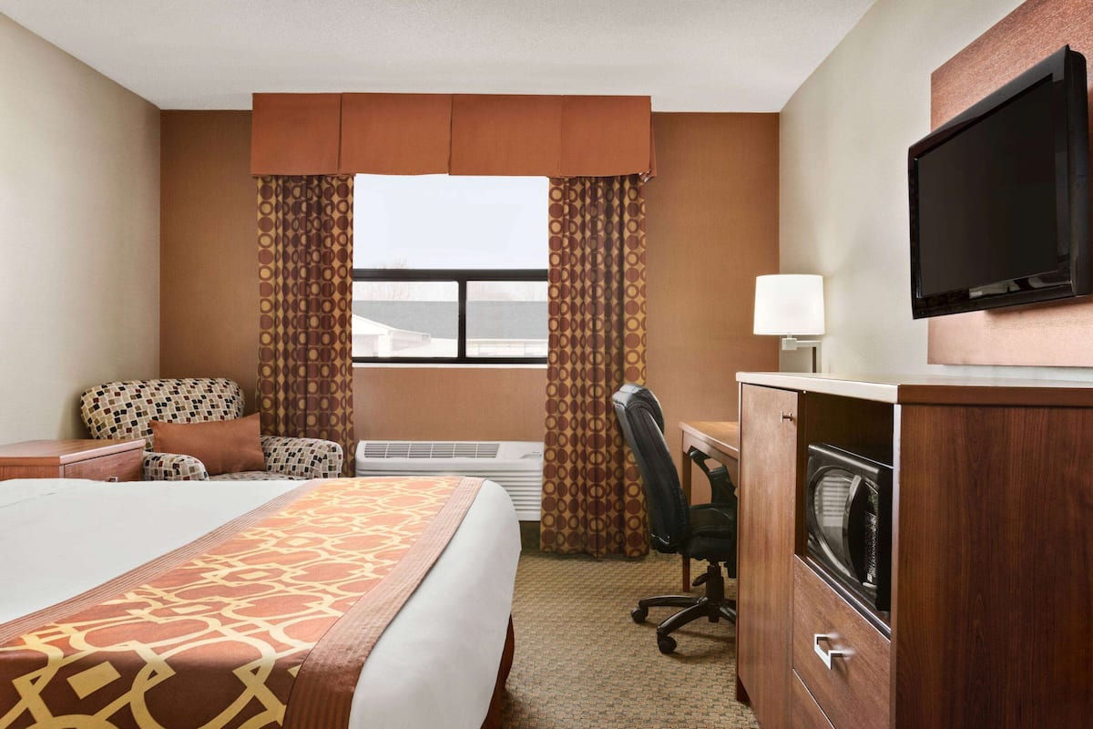 A hotel room with a bed, a desk with a television, a chair, an air conditioning unit, and curtains.