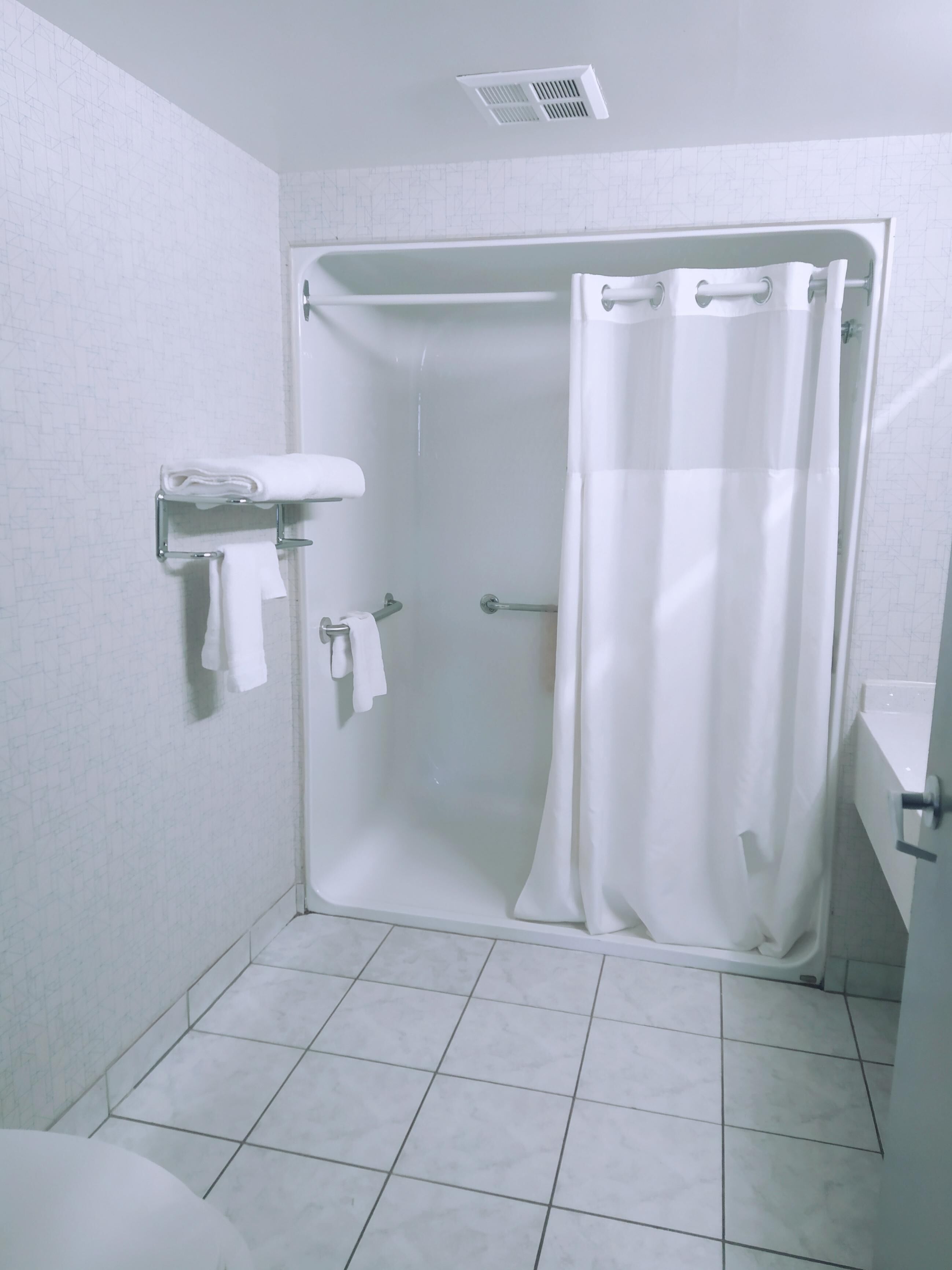 A shower with a white curtain, a towel rack with towels, and a toilet visible in a bathroom with white tiled walls and floor.