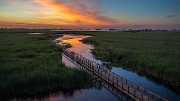 A wooden bridge over a calm water body with grassy banks, set against a sunset sky.