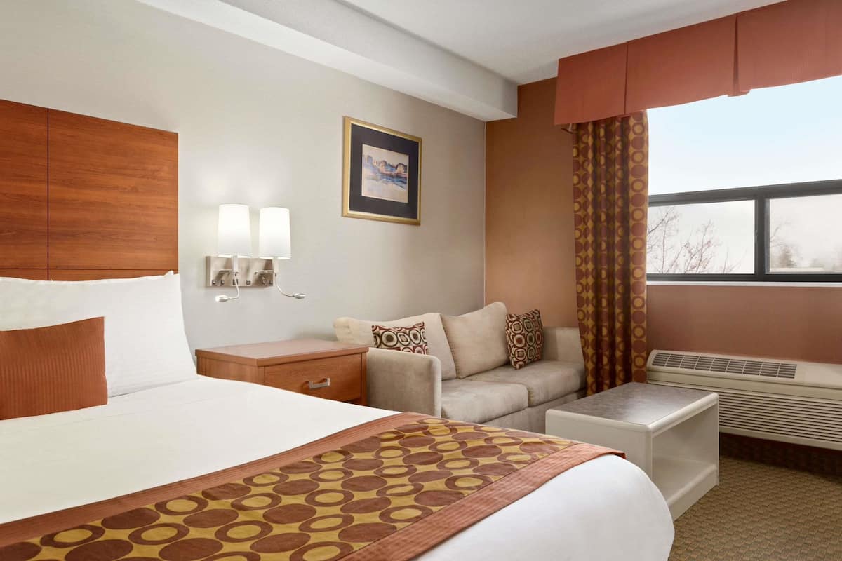 A hotel room with a large bed, a sofa, a small table, a nightstand, a wall-mounted lamp, a picture, and a window with curtains.