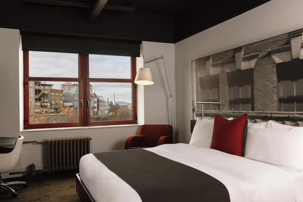 A hotel room with a large bed, a desk, a chair, a radiator, and a window with a city view.