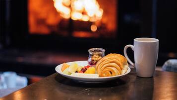 Un plato con croissant, fruta y un vaso pequeño de mermelada sobre una mesa con una taza blanca, frente a una chimenea.