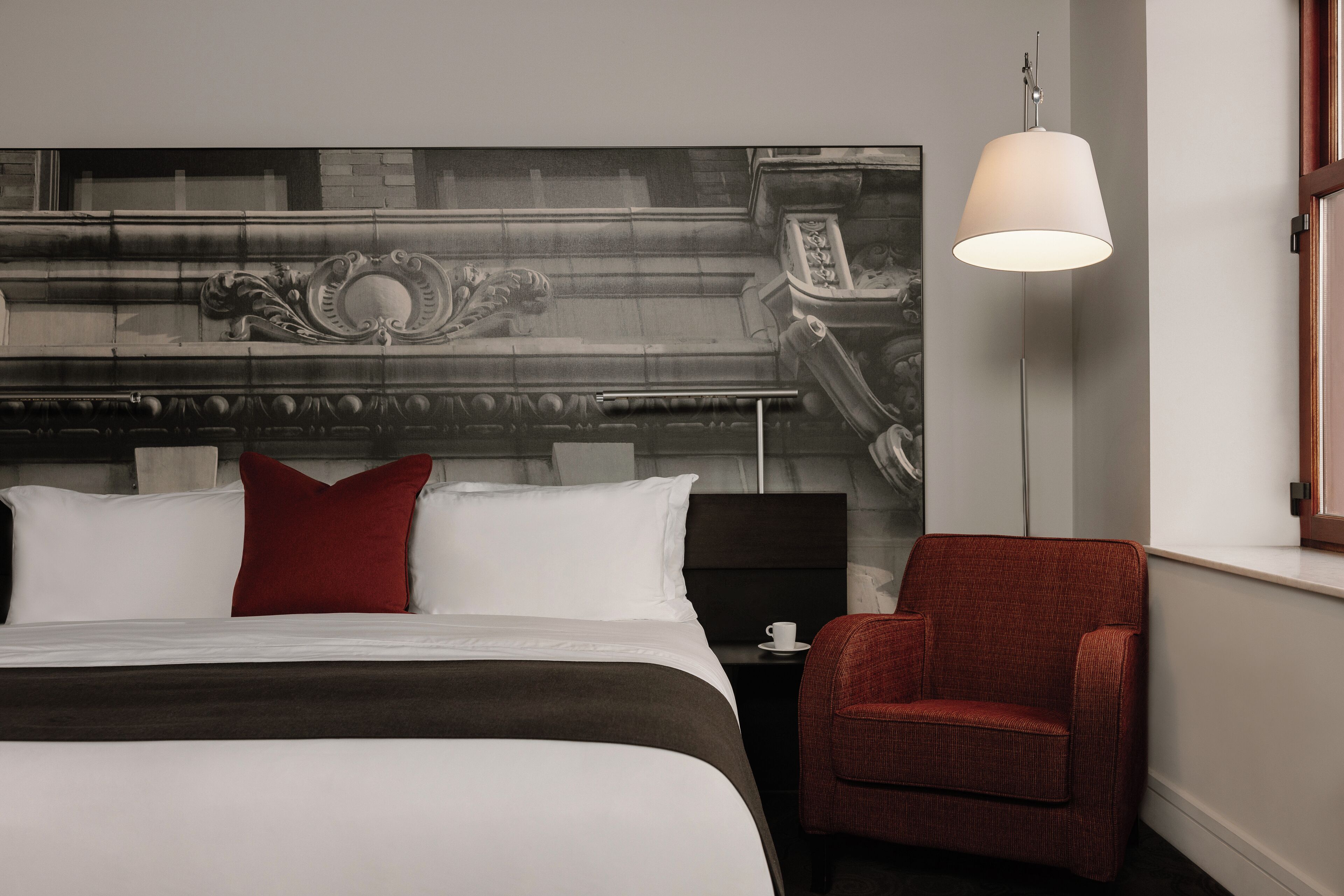 Habitación de hotel con una cama grande, una silla roja, una mesita de noche con una taza y una lámpara de pared.