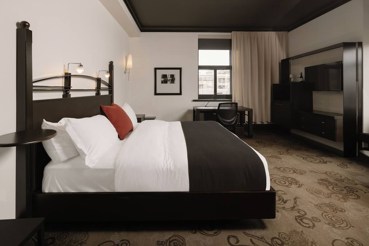 A hotel room with a large bed, a desk, a chair, a TV, and a window with curtains.
