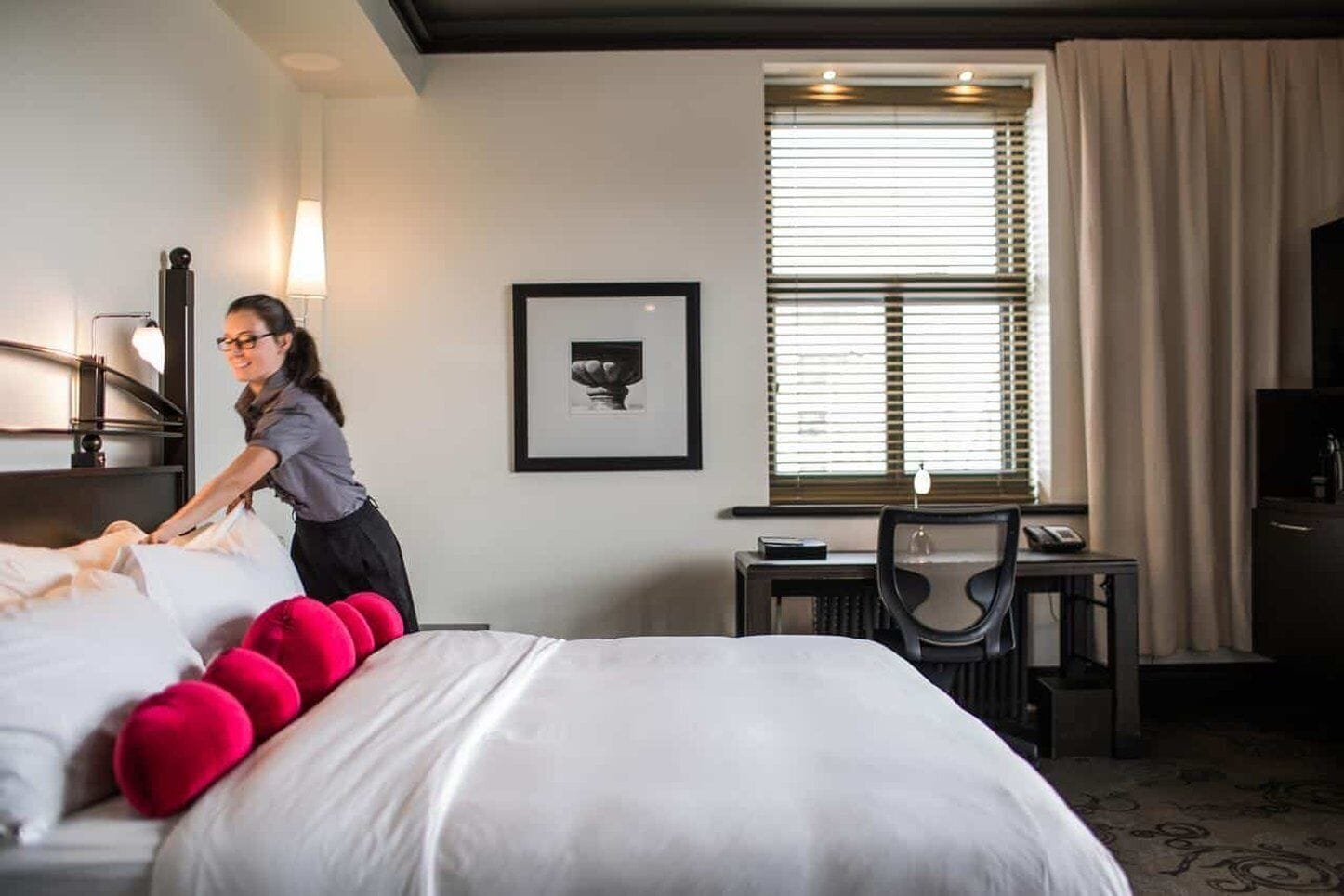 A hotel room with a neatly made bed, a desk with a chair, a framed picture on the wall, and a window with blinds.