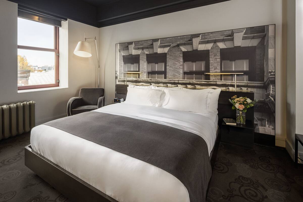 A modern hotel room with a large bed, a headboard with a stone-like design, a side table with a vase of flowers, and a chair.